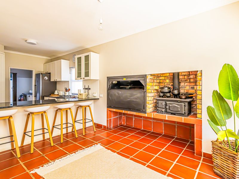 Open-plan kitchen and living area featuring a built-in braai, cast iron stove, terracotta tiled flooring, and a functional breakfast bar with granite countertops.