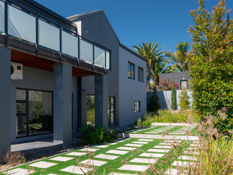 Rear view of a modern two-story house featuring a balcony with glass balustrades, a paved patio area, and a well-maintained landscaped garden.