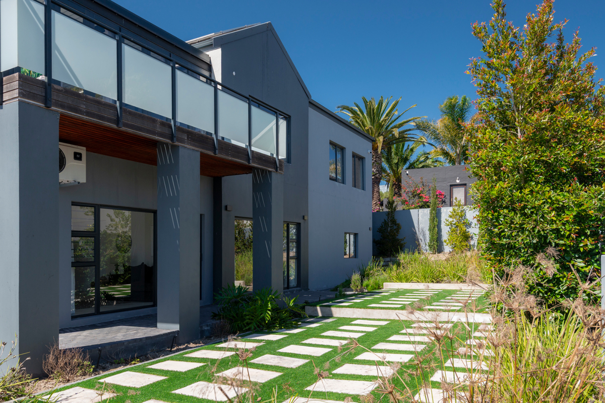 Rear view of a modern two-story house featuring a balcony with glass balustrades, a paved patio area, and a well-maintained landscaped garden.