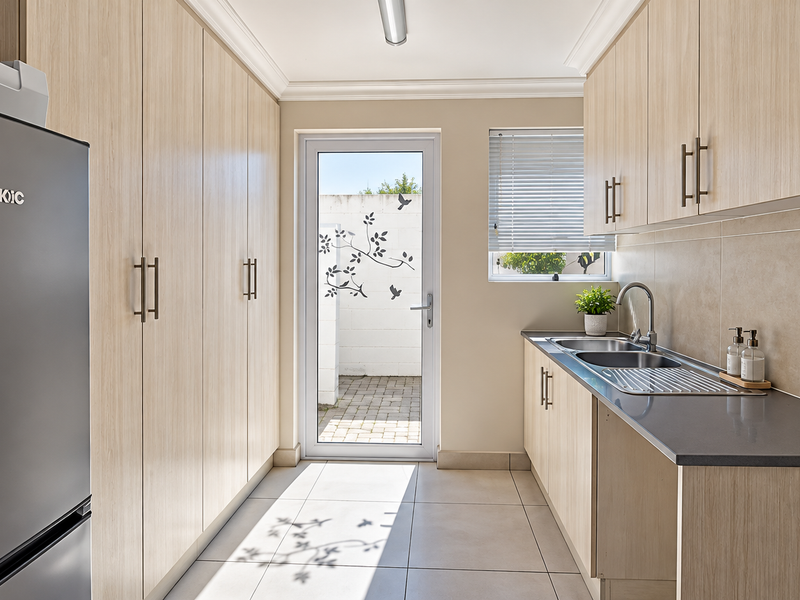A modern, well-maintained scullery featuring extensive built-in cabinetry, a stainless steel sink, and direct access to an outdoor paved area.