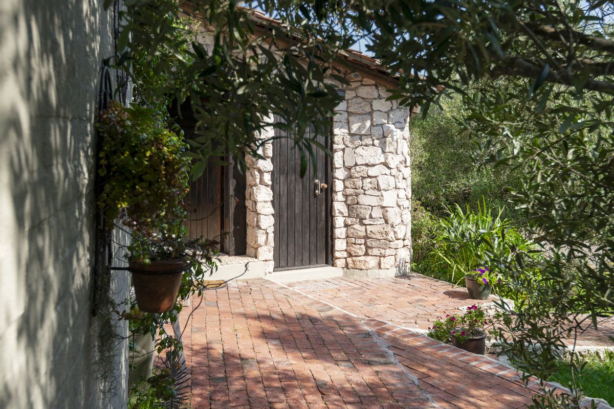A charming rear exterior view featuring rustic stone masonry walls, a solid timber door, and traditional brick paving surrounded by lush garden greenery.
