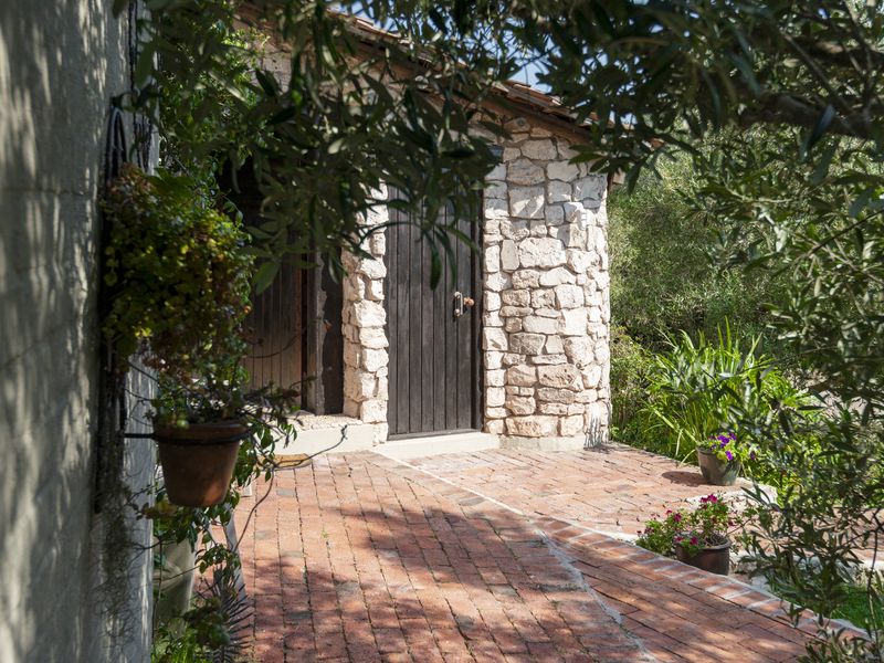 A charming rear exterior view featuring rustic stone masonry walls, a solid timber door, and traditional brick paving surrounded by lush garden greenery.