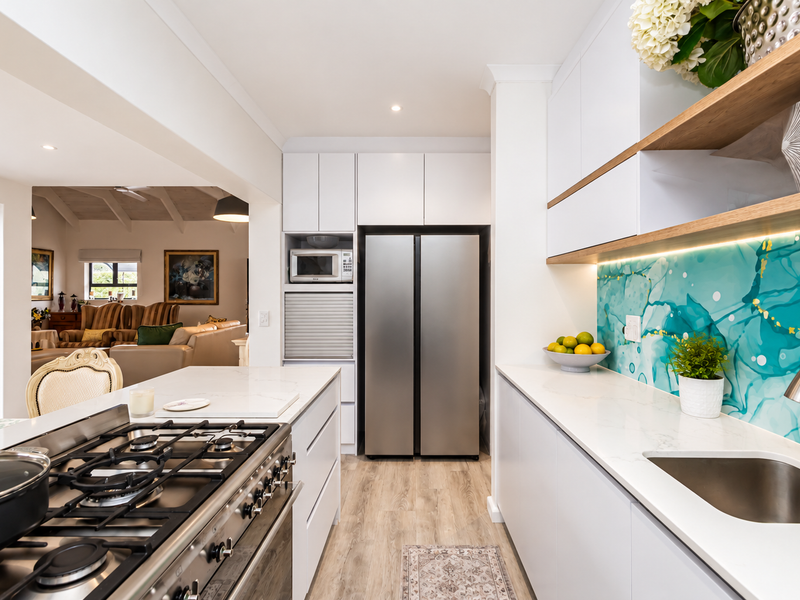 Modern kitchen featuring white cabinetry, stone countertops, a gas cooktop, built-in appliances, and a vibrant feature splashback, seamlessly flowing into the living area.