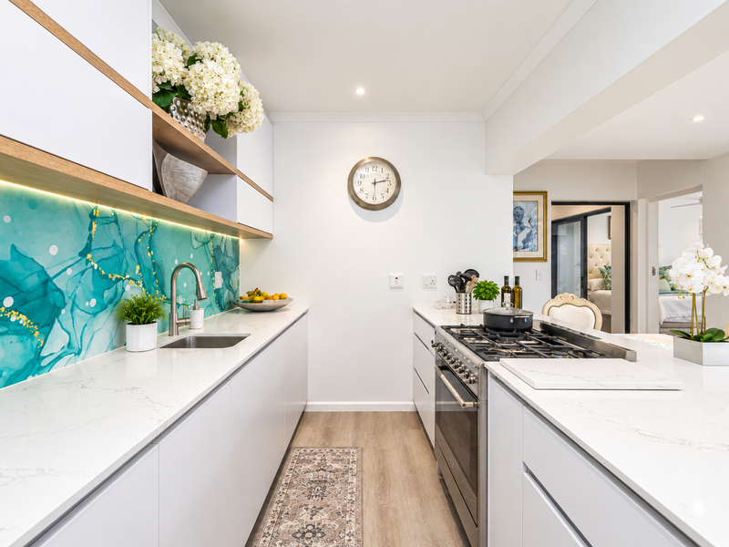 Modern galley-style kitchen featuring white cabinetry, quartz countertops, a striking teal glass splashback, and a high-end stainless steel gas range.