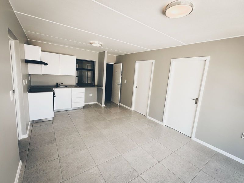 A bright, open-plan living and kitchen area featuring neutral grey walls, modern tiled flooring, and clean white cabinetry with integrated fixtures.