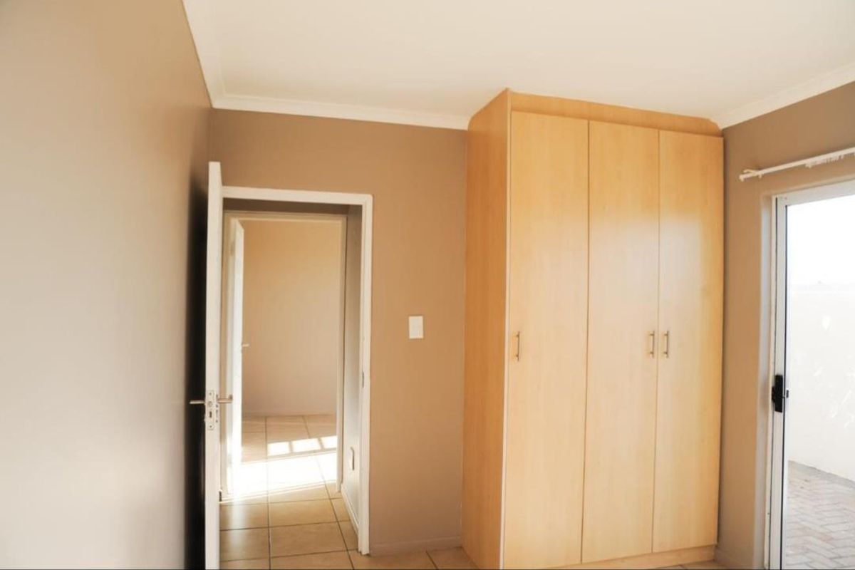 A clean, empty bedroom featuring neutral wall tones, built-in wooden wardrobes, tiled flooring, and direct access to an outdoor area via a sliding glass door.