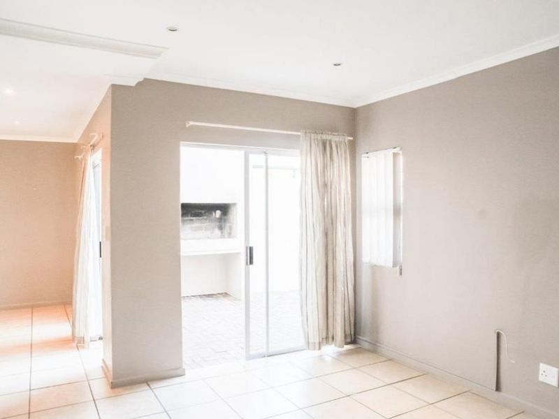 An empty living area featuring neutral tiled flooring, coved ceilings, and a sliding glass door providing access to an outdoor patio with a built-in braai.