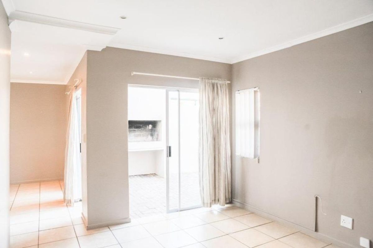 An empty living area featuring neutral tiled flooring, coved ceilings, and a sliding glass door providing access to an outdoor patio with a built-in braai.