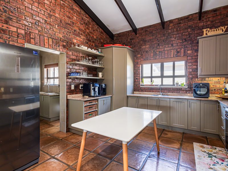 A rustic kitchen featuring exposed brick walls, timber ceiling beams, fitted cabinetry, stone countertops, and a tiled floor, with an adjacent scullery area.