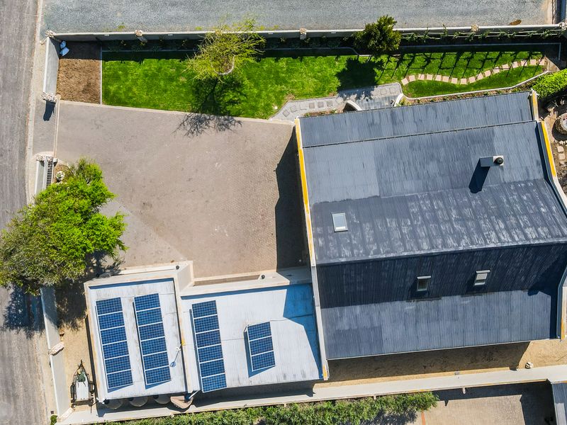 Aerial view of the property showing the main house with solar panels, corrugated metal roofing, skylights, a paved driveway, and a landscaped garden.