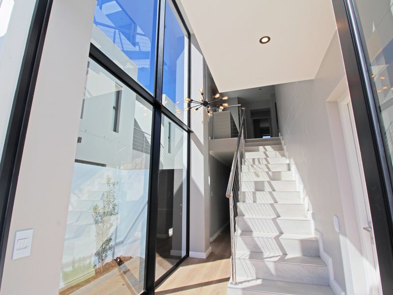 Modern double-volume entrance hall featuring floor-to-ceiling glass panels, timber flooring, and a sleek staircase with stainless steel balustrades.