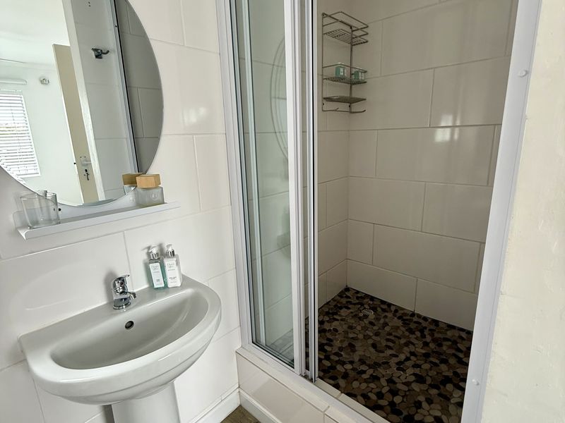 A clean, modern bathroom featuring a pedestal washbasin, a circular wall mirror, and a shower enclosure with a distinctive pebble-tiled floor.