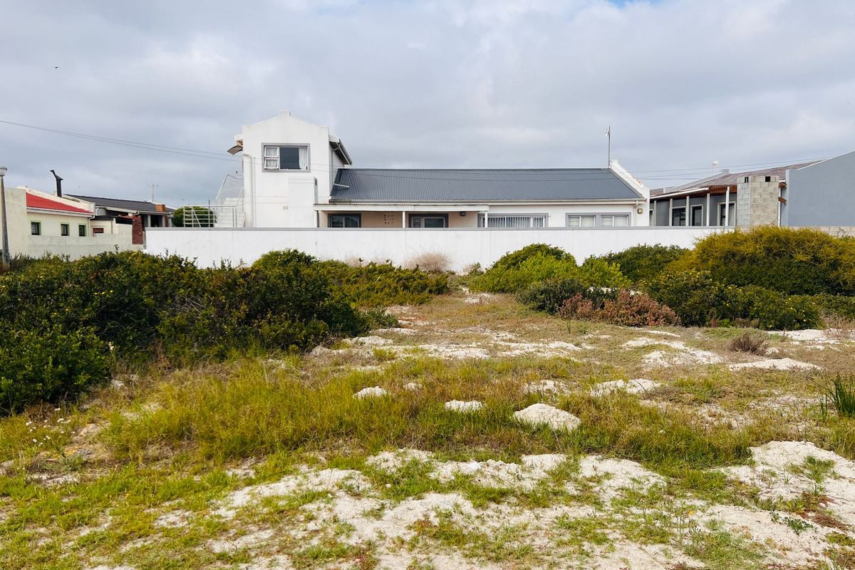 A view of a vacant residential plot featuring natural coastal vegetation, with a neighboring house visible in the background.
