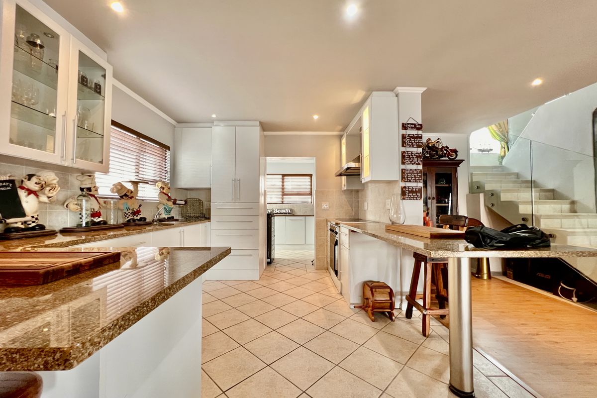 A well-lit kitchen featuring granite countertops, white cabinetry, tiled flooring, and an integrated breakfast bar, with a staircase visible in the background.
