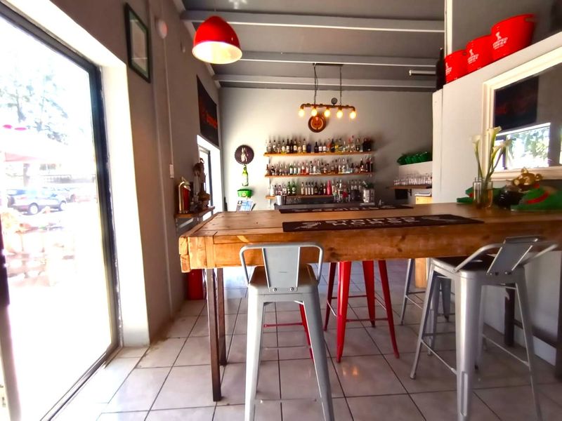 A well-lit bar area featuring a wooden counter, wall-mounted shelving for display, tiled flooring, and an exposed beam ceiling with direct access to the exterior.