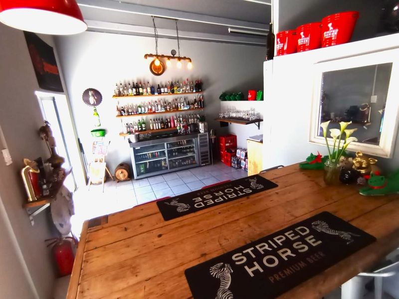 A functional bar area featuring a wooden counter, under-counter refrigeration, wall-mounted shelving for glassware and spirits, and industrial-style lighting.