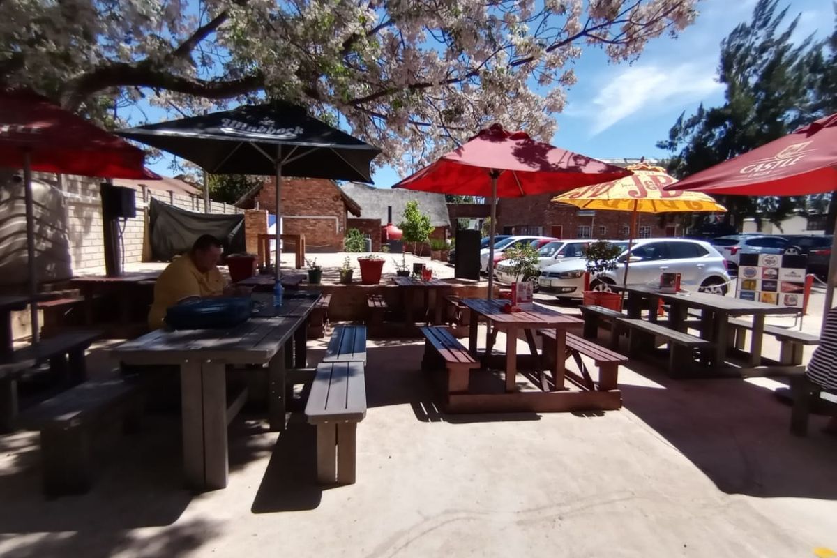 Outdoor patio area featuring paved flooring and brick boundary walls, shaded by mature trees and large umbrellas, suitable for casual dining or social gatherings.