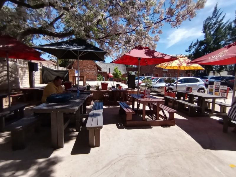 Outdoor patio area featuring paved flooring and brick boundary walls, shaded by mature trees and large umbrellas, suitable for casual dining or social gatherings.