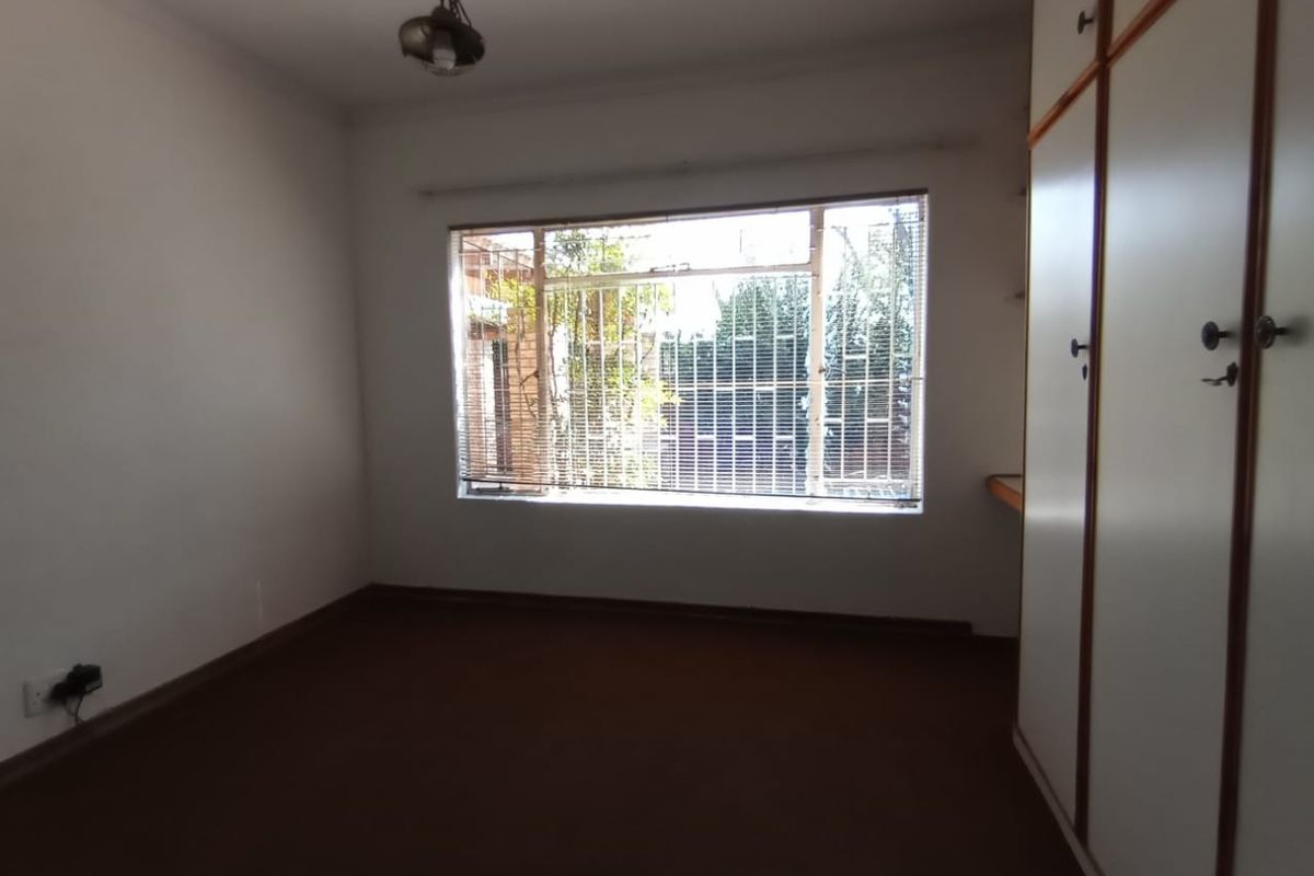 A vacant bedroom featuring built-in cupboards, carpeted flooring, and a large window fitted with security bars.