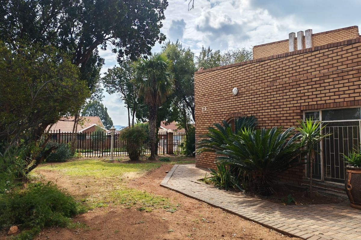 A rear exterior view of a face brick house featuring a paved walkway, established garden, and secure metal perimeter fencing.