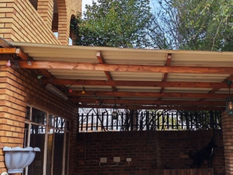 A covered outdoor patio area featuring brick construction, timber roofing beams, and a built-in brick braai, providing a functional space for outdoor entertaining.