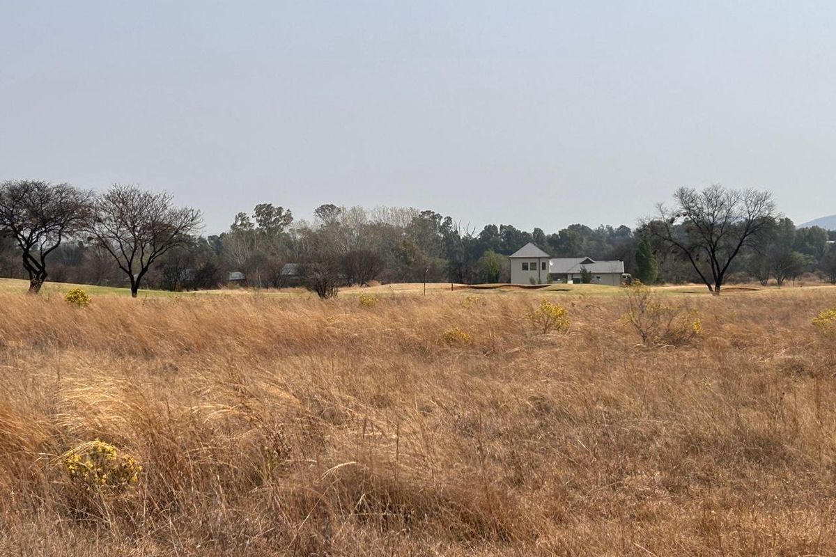 A view of an expansive, dry grassland plot with mature trees and distant residential structures in the background.