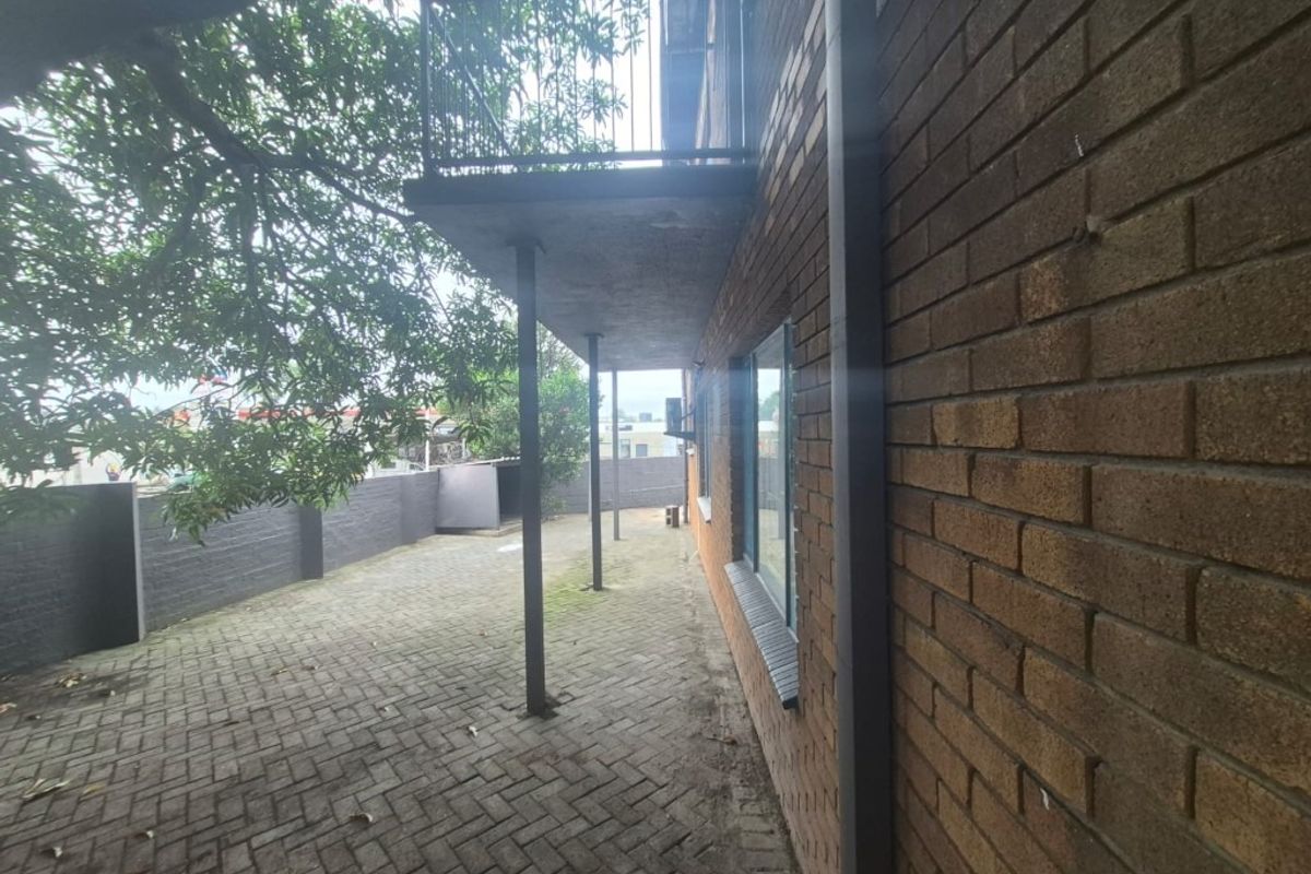Rear exterior view of the flat featuring a paved courtyard area, brick facade, and an overhead balcony with metal railings.