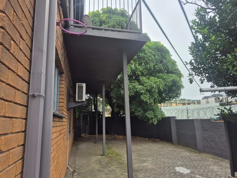 A covered ground-level patio area featuring brick walling, paved flooring, and an overhead balcony structure with metal railings.
