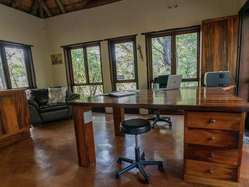 A spacious home office featuring a high thatched ceiling, polished concrete floors, and extensive built-in wooden cabinetry with large windows overlooking the garden.
