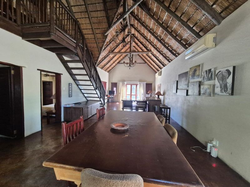 Spacious open-plan living and dining area featuring a high thatched roof with exposed timber trusses, a wooden staircase, and polished concrete flooring.