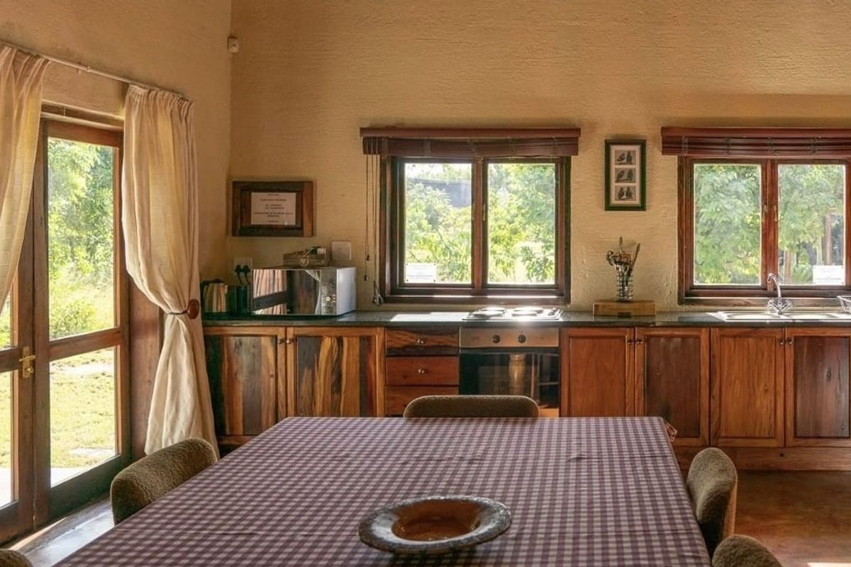 A rustic kitchen featuring wooden cabinetry, stone countertops, built-in oven and hob, and large timber-framed windows providing natural light and garden views.