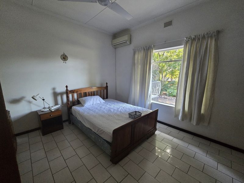 A functional bedroom featuring tiled flooring, a ceiling fan, and a wall-mounted split-system air conditioner with natural light from a large window.