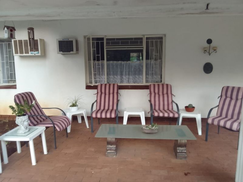 A covered patio area featuring brick flooring, a large window with security bars, and a wall-mounted air conditioning unit.