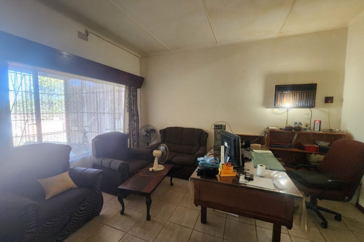 A living room area featuring tiled flooring, large windows with security bars, and a ceiling fan, currently utilized as a multi-purpose living and workspace.