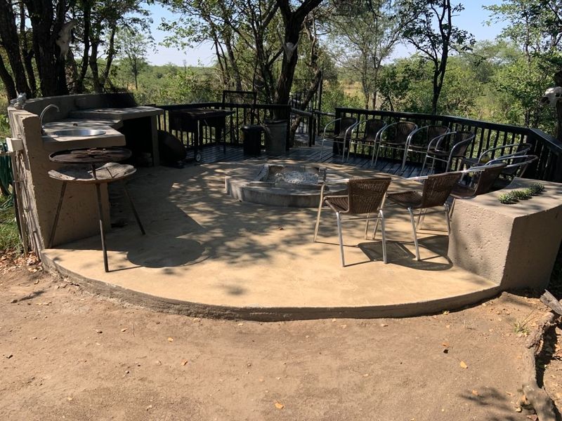 Outdoor entertainment area featuring a built-in braai, concrete prep station with sink, central fire pit, and timber decking overlooking natural surroundings.