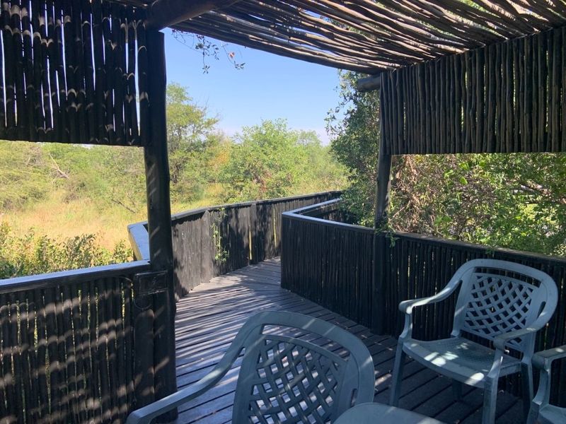 An elevated timber deck patio featuring a rustic timber pole structure and slatted roof, overlooking a natural bushveld landscape.