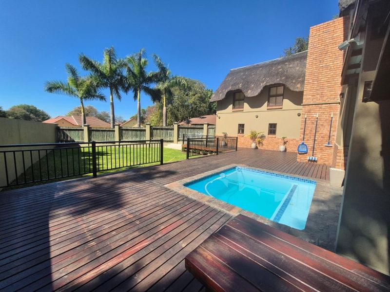 A well-maintained outdoor pool area featuring extensive timber decking, a brick chimney, and a thatched roof house exterior with a manicured lawn.