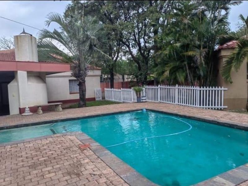 Outdoor pool area featuring brick paving, a built-in fireplace, and a white picket fence, set within a landscaped garden environment.