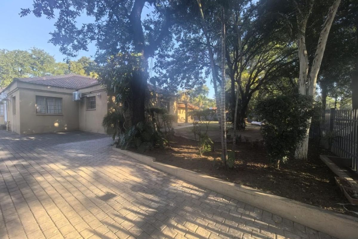 A view of the rear exterior featuring a paved driveway, mature trees, and a well-established garden area adjacent to the main house structure.