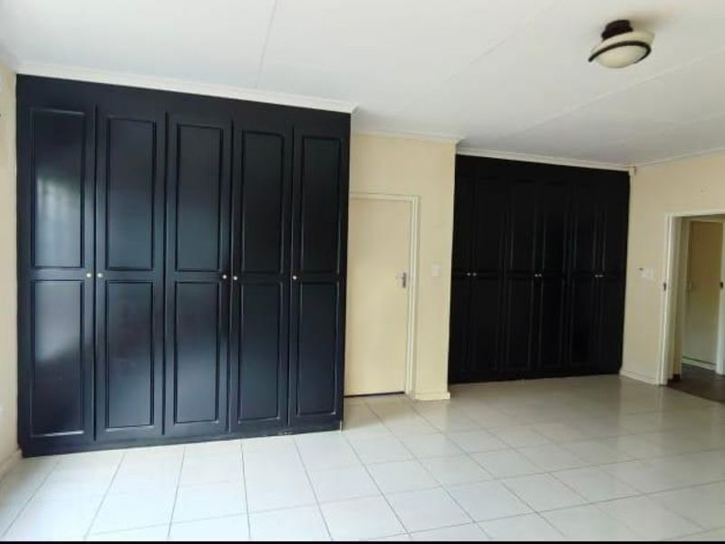 A spacious bedroom featuring extensive dark-toned built-in cupboards, neutral tiled flooring, and a simple ceiling light fixture.