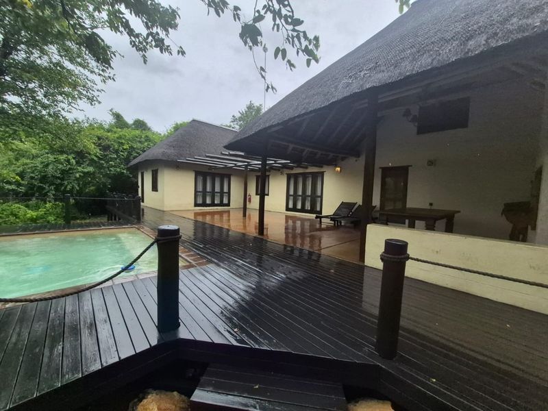 Outdoor pool area featuring extensive timber decking, a covered patio space, and a thatched roof structure set within a lush garden environment.