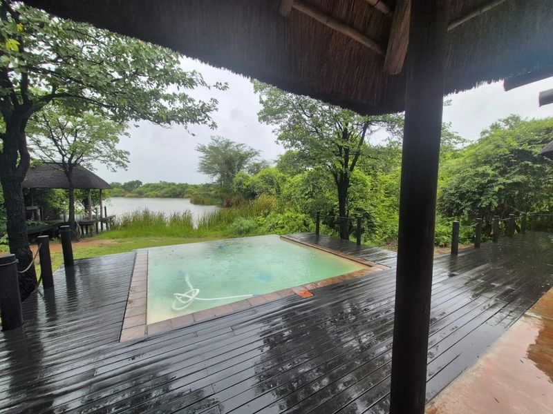 Outdoor pool area featuring timber decking and a thatched roof structure, overlooking a natural lake and lush greenery.