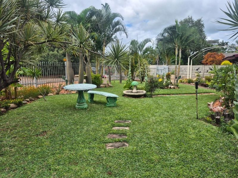 A well-maintained garden featuring lush lawn, mature palm trees, stone pathways, and a decorative concrete water feature enclosed by steel perimeter fencing.