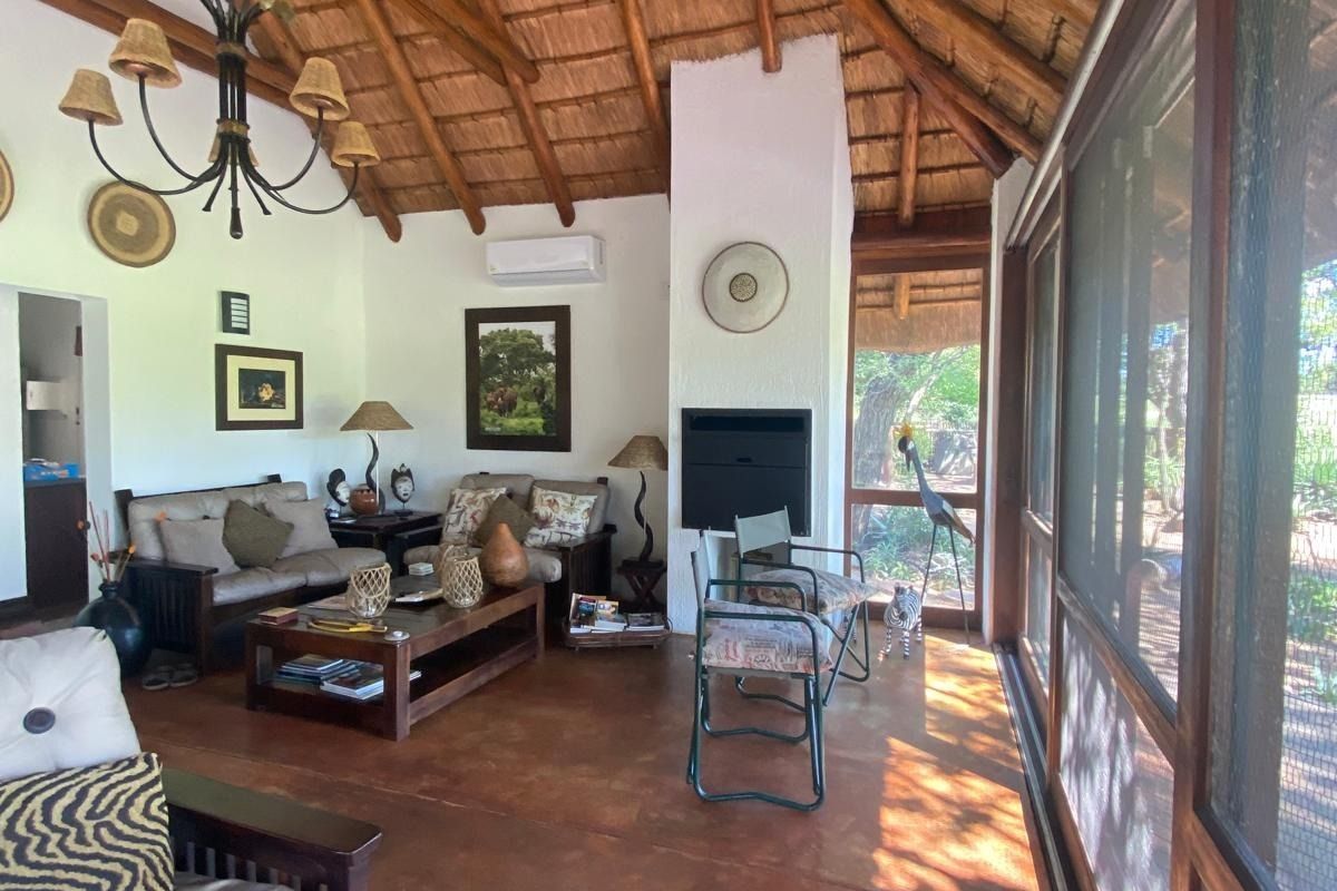 Spacious living area featuring a high thatched ceiling with exposed timber rafters, a built-in fireplace, and large timber-framed screen windows overlooking the garden.