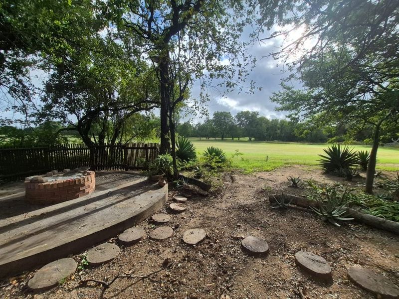 A spacious garden area featuring a circular brick fire pit, concrete patio space, and natural landscaping with mature trees and a wooden fence.