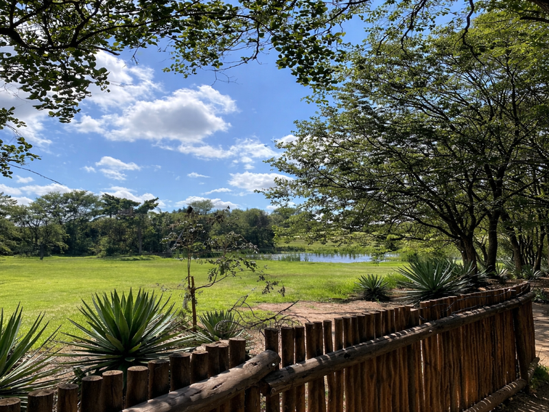 A scenic view of a lush green lawn, mature trees, and a natural pond, framed by a rustic wooden pole fence.