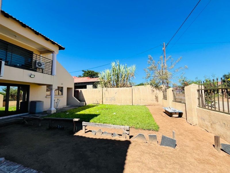 Rear garden area featuring a lawn, boundary wall with security fencing, and a two-story house structure with an upper-level balcony.