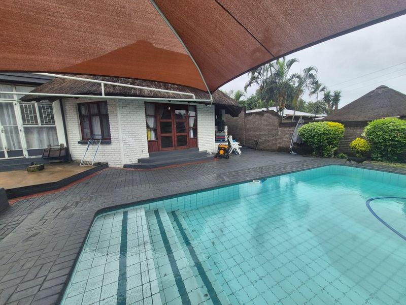 Outdoor pool area featuring a swimming pool, brick paving, and a large shade sail, adjacent to a house with a traditional thatched roof.