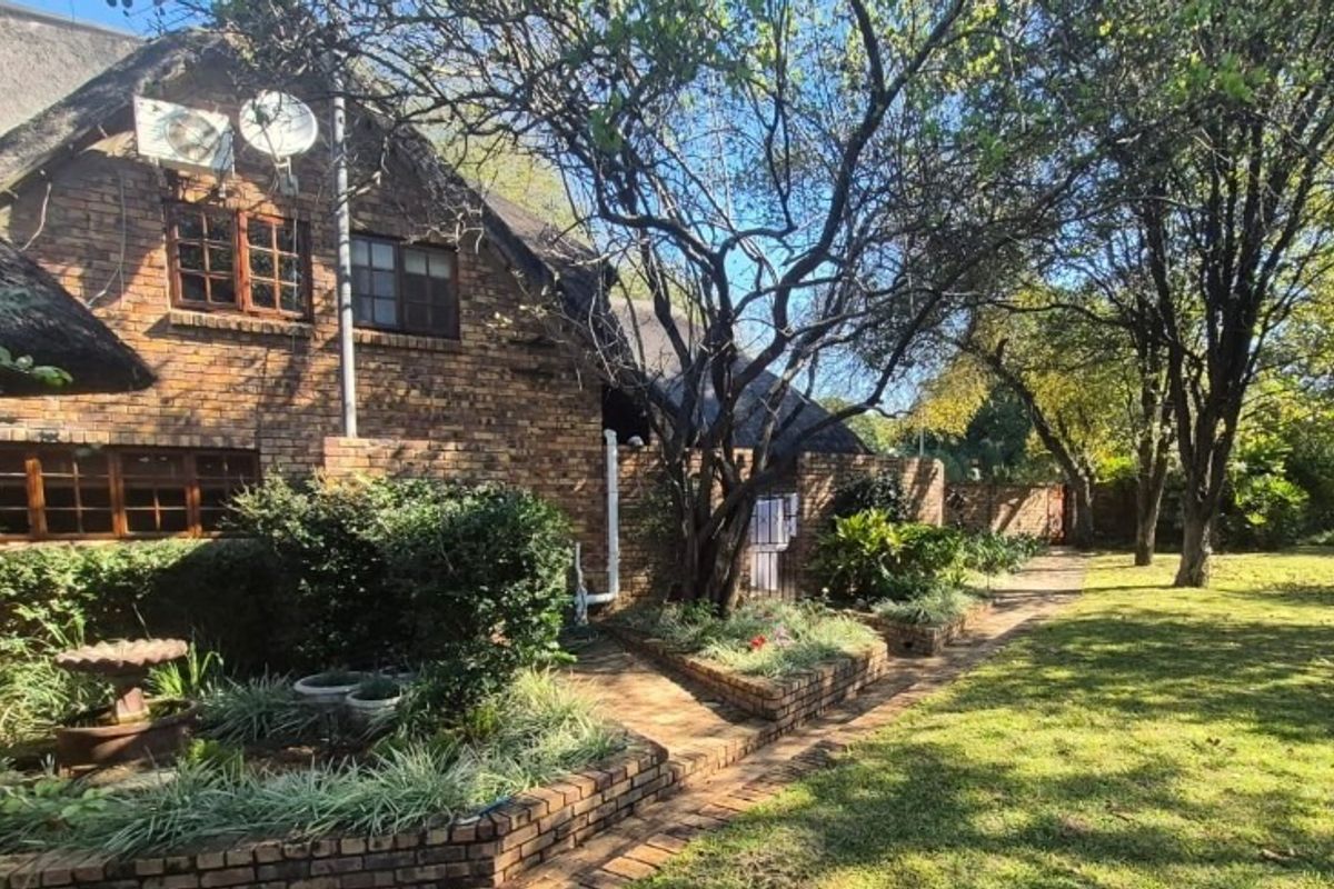 Rear view of a brick house with a thatched roof, featuring established garden beds, paved pathways, and mature trees in a spacious backyard setting.