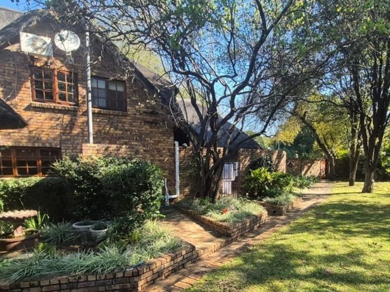 Rear view of a brick house with a thatched roof, featuring established garden beds, paved pathways, and mature trees in a spacious backyard setting.